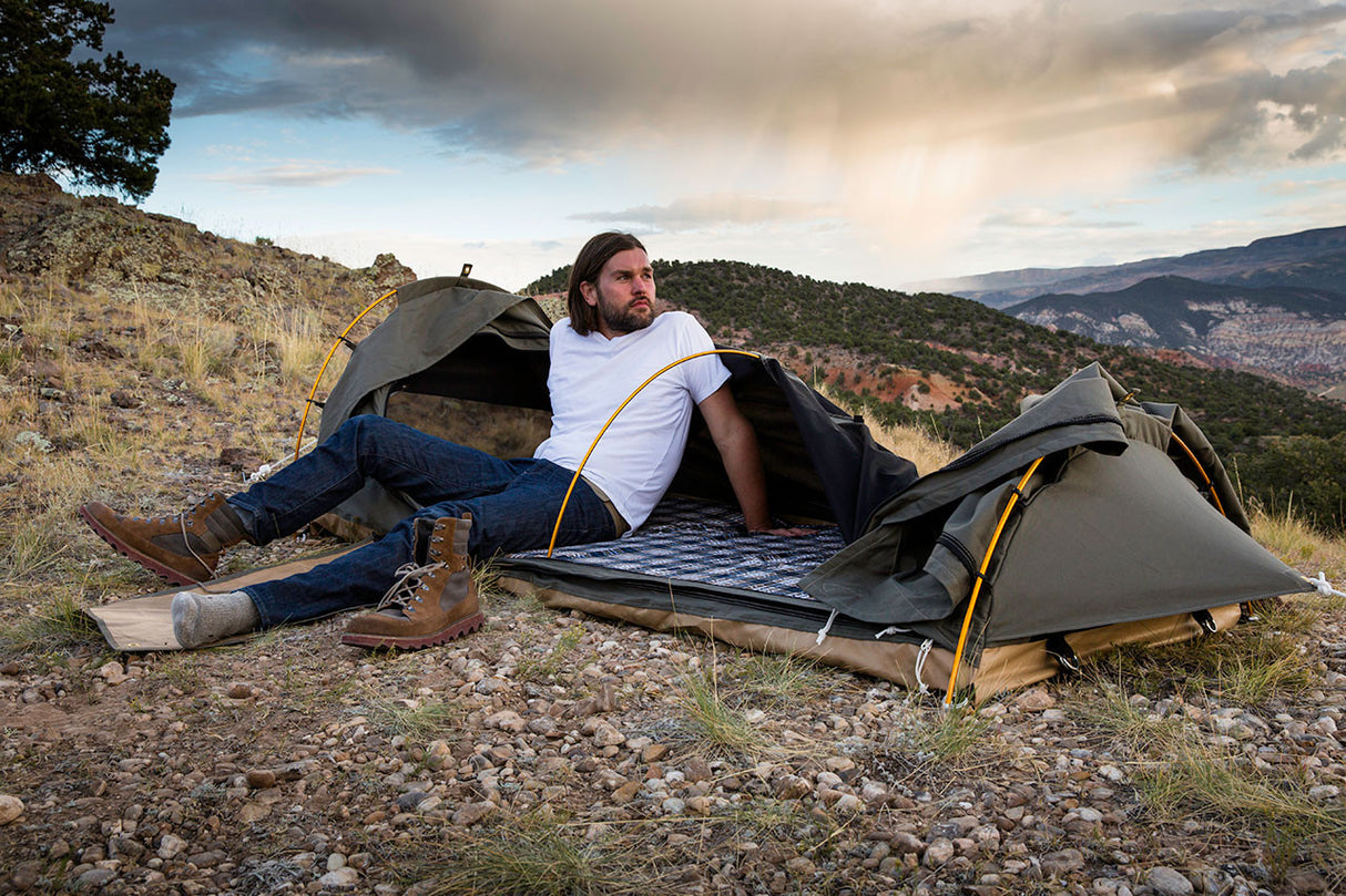 Outdoor camping tent in olive green, featuring a man relaxing inside with rugged terrain in background.