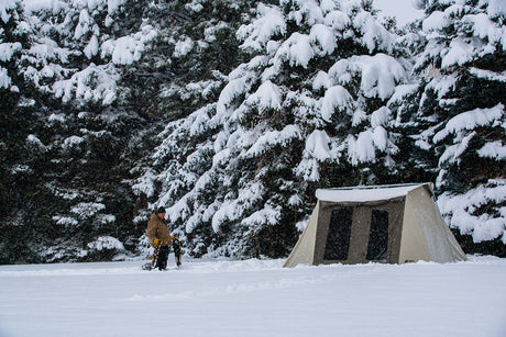Cozy winter camping tent in snow, beige canvas, with large mesh windows, surrounded by snow-covered trees.