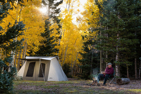 Large beige camping tent set in a vibrant autumn forest, with a man relaxing by a campfire.