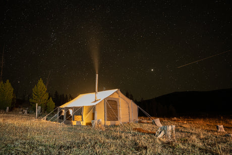 Canvas tent with chimney at night, surrounded by stars and trees, ideal for camping and glamping.