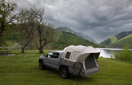 Gray pickup truck with beige rooftop tent by a lake, surrounded by lush green mountains.