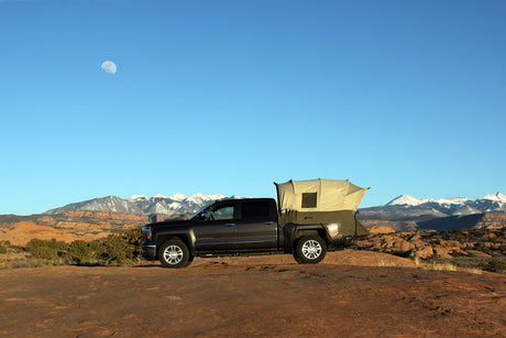 Gray pickup truck with a tan rooftop tent set against a mountain backdrop and clear blue sky.