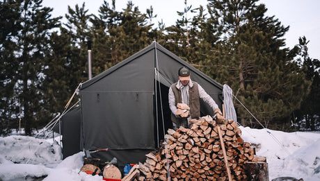 Man stacking firewood in front of a green camping tent surrounded by snow and trees.