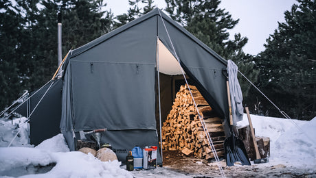 Spacious gray canvas camping tent with firewood stack, snowy ground, and outdoor gear.