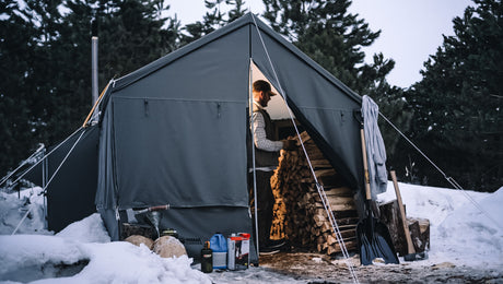 Grey canvas tent in snowy forest with firewood and camping gear outside.