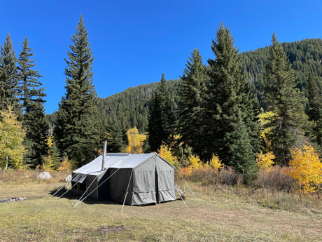 Durable gray canvas camping tent with stove pipe, set in scenic forested area, perfect for outdoor adventures.