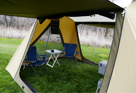 Cozy camping tent with tan fabric, two blue chairs, and a white folding table on green grass.